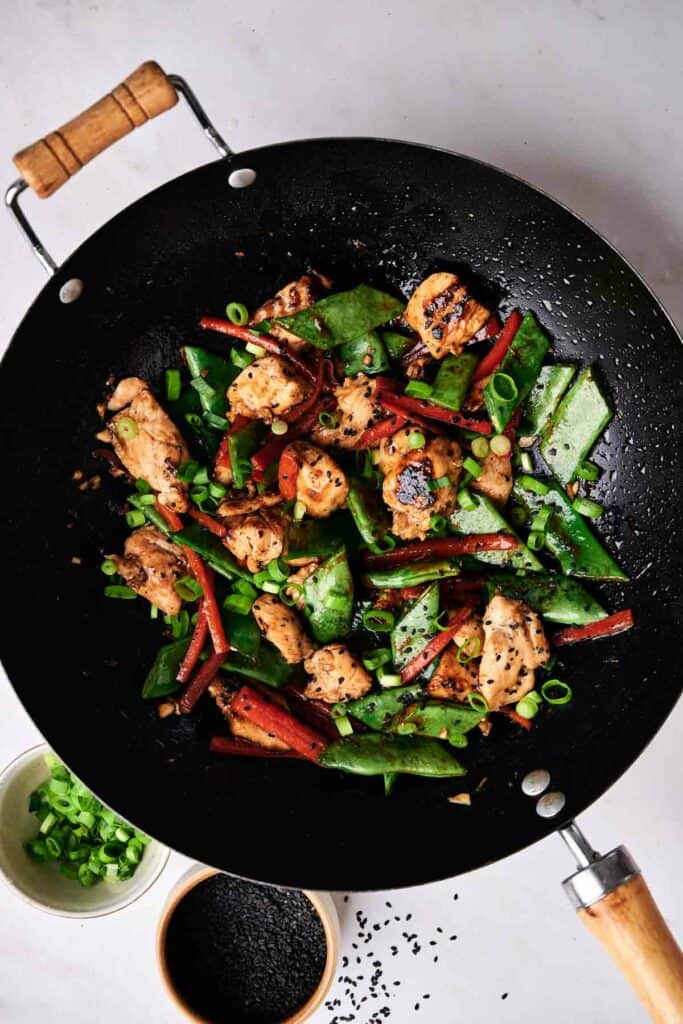 A wok filled with a colorful chicken and vegetable stir-fry similar to kung pao chicken, featuring snow peas, red bell peppers, and garnished with chopped green onions and black sesame seeds. Two small bowls with green onions and sesame seeds are nearby.