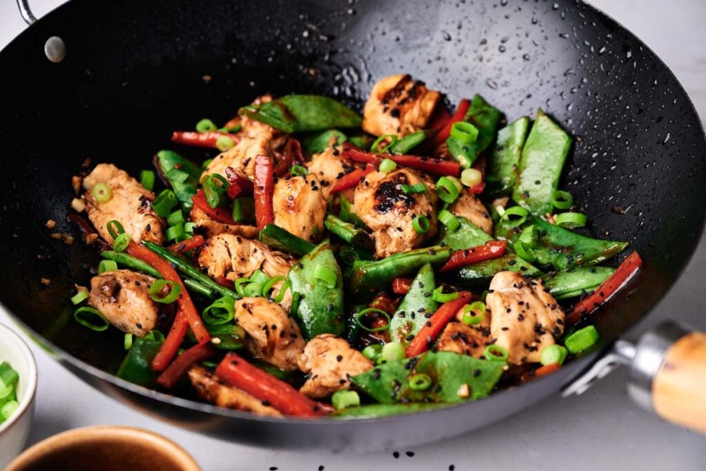 Kung pao chicken stir-fry with snow peas, carrots, green onions, and black sesame seeds cooking in a black wok.