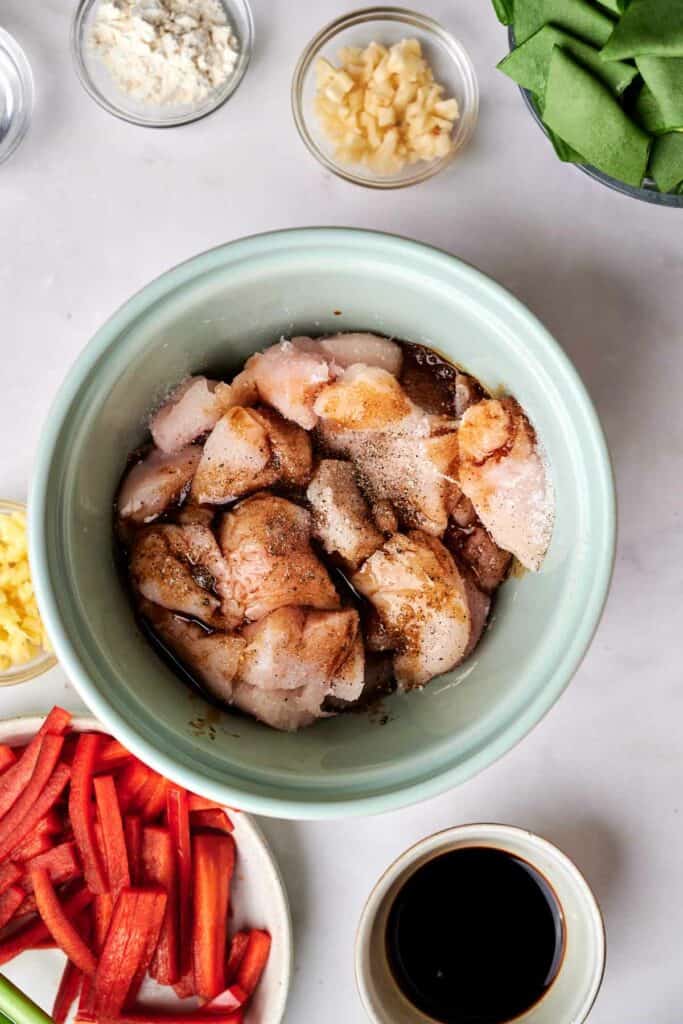 A bowl of raw chicken pieces marinated in seasoning, perfect for kung pao chicken, surrounded by small bowls of chopped garlic, diced ginger, red bell peppers, green vegetables, flour, and a small bowl of soy sauce.