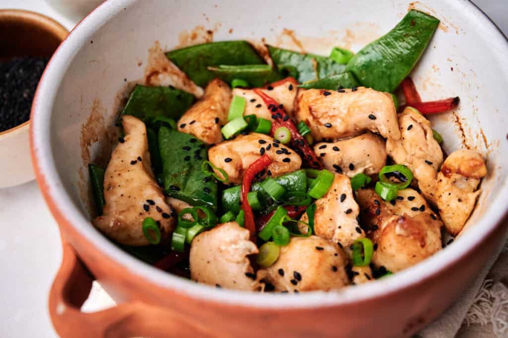 A bowl of kung pao chicken with snap peas, bell peppers, scallions, and sesame seeds.