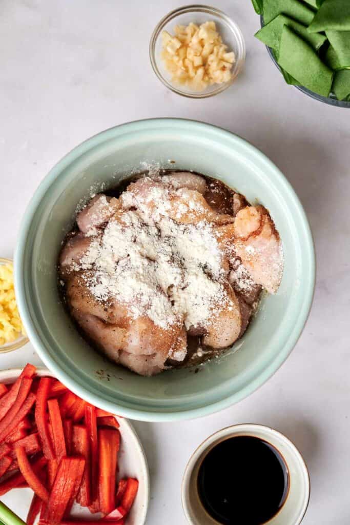 A bowl of raw, seasoned chicken pieces with flour is surrounded by ingredients like cut red bell peppers, soy sauce, garlic, and other vegetables&mdash;perfect for making a flavorful kung pao chicken.