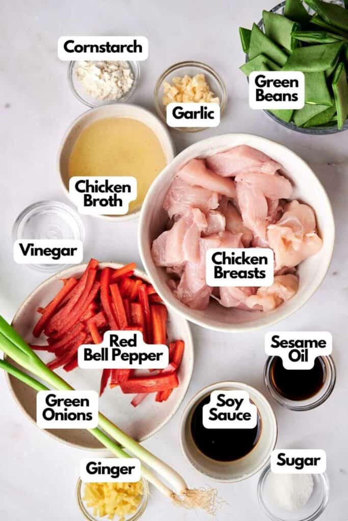 Ingredients for a stir-fry recipe displayed on a white surface. Includes chicken breasts, green beans, red bell pepper, green onions, garlic, ginger, cornstarch, vinegar, soy sauce, sesame oil, chicken broth, and sugar&mdash;perfect essentials to craft a delicious Kung Pao Chicken.
