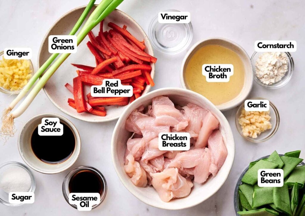 Bowls and dishes containing kung pao chicken stir-fry ingredients: green onions, ginger, soy sauce, sugar, red bell pepper, sesame oil, chicken breasts, vinegar, chicken broth, garlic, cornstarch, and green beans.