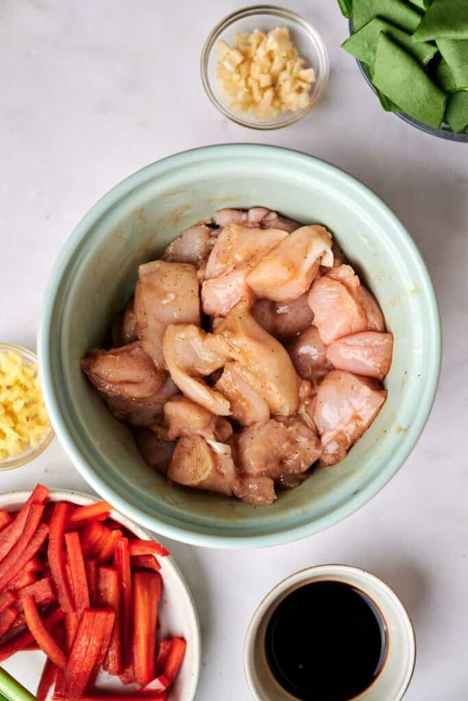 A bowl of raw, marinated chicken pieces is surrounded by bowls containing chopped garlic, chopped ginger, sliced red bell peppers, green vegetables, and a small bowl of sauce on a white surface&mdash;perfect for crafting a delicious kung pao chicken.