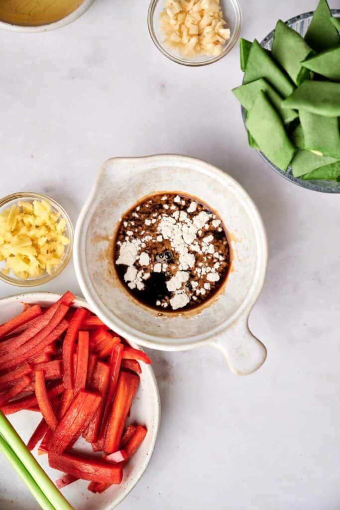 A white bowl with sauce and flour mixture, surrounded by small bowls of chopped garlic, ginger, and green vegetables, as well as a plate of sliced red bell peppers on a light-colored surface&mdash;perfect for preparing kung pao chicken.