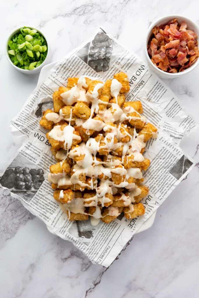 Plate of loaded tater tots with drizzled sauce, bacon bits, and chopped green onions, served on a newspaper-lined tray on a marble surface.