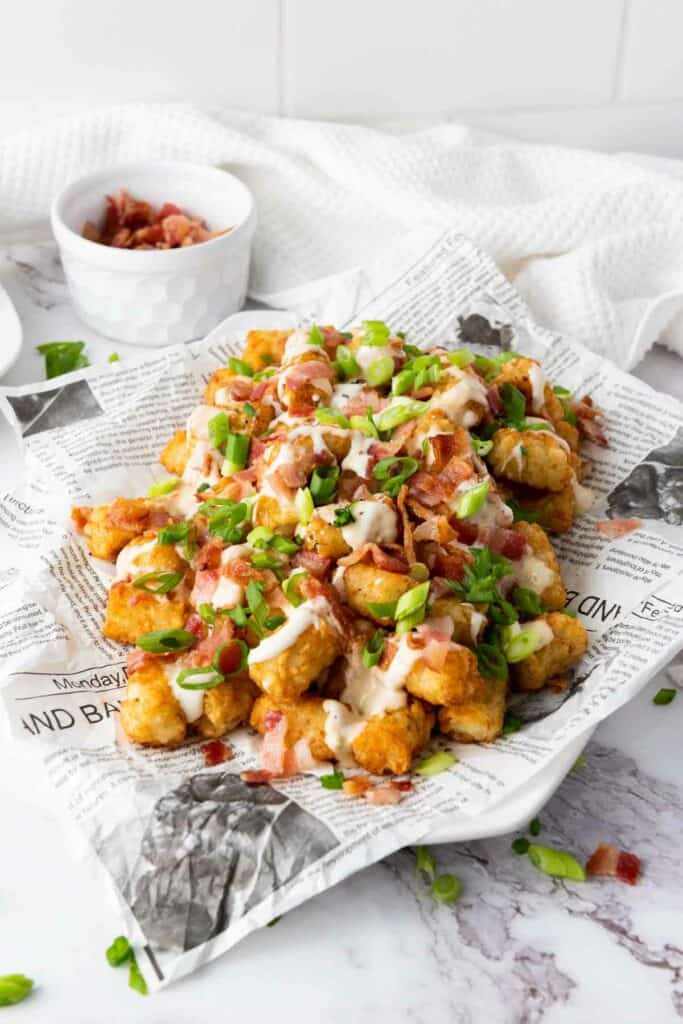  A plate of loaded tater tots topped with melted cheese, bacon bits, and green onions, served on a newspaper-lined plate.