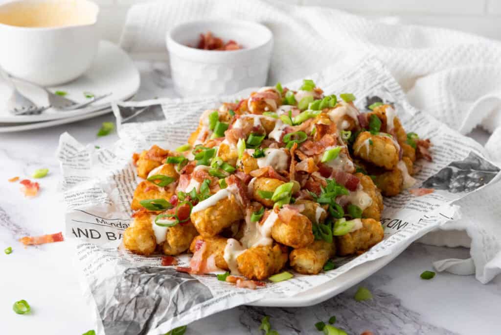 A plate of Loaded Tater Tots topped with bacon, green onions, and drizzled with cheese sauce, served on a newspaper-lined tray.