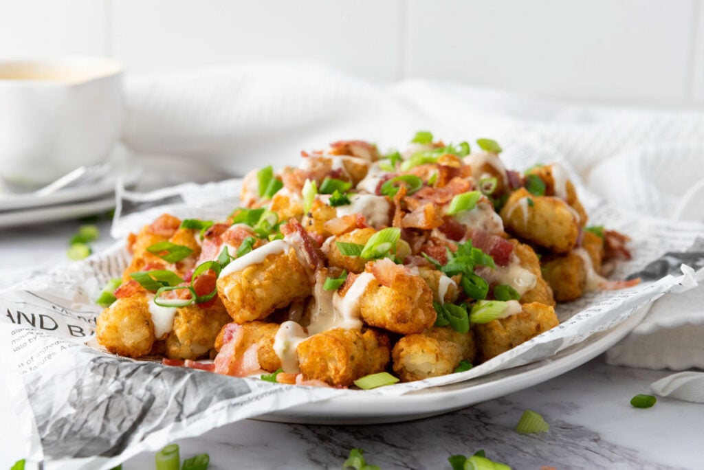 A plate of Loaded Tater Tots topped with chopped bacon, green onions, and a creamy sauce, served on newspaper-style paper.