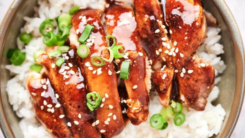 A bowl of rice topped with glazed chicken, garnished with sesame seeds and sliced green onions.