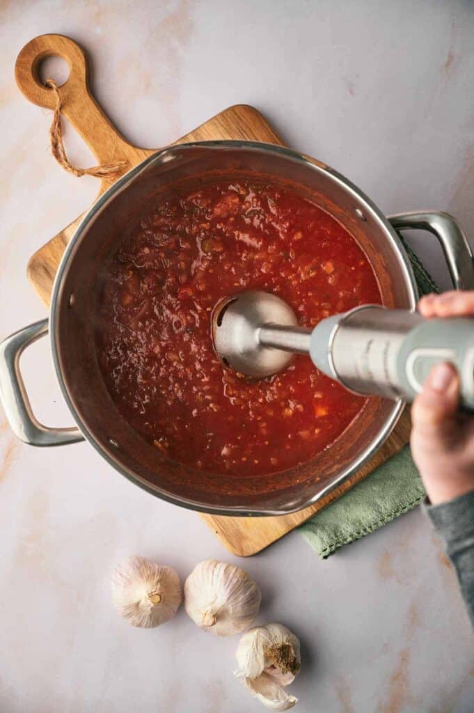 A person blending tomato sauce in a pot with an immersion blender, garlic bulbs nearby on a kitchen counter.