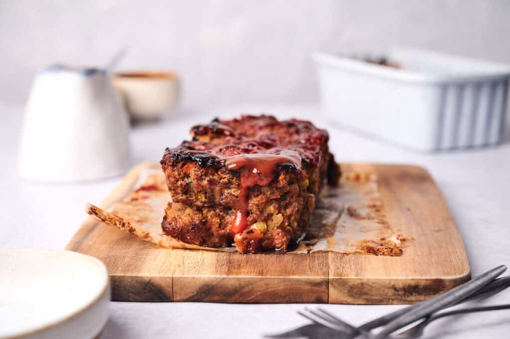 A succulent sliced meatloaf topped with rich sauce rests on a wooden cutting board, with two forks and a pristine white dish in the background.