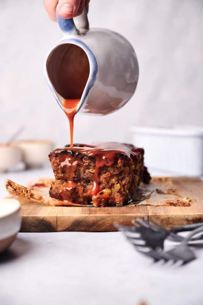 Hand pouring sauce over a slice of meatloaf on a wooden cutting board, with dishes and cutlery in the background, enhancing the classic comfort food.