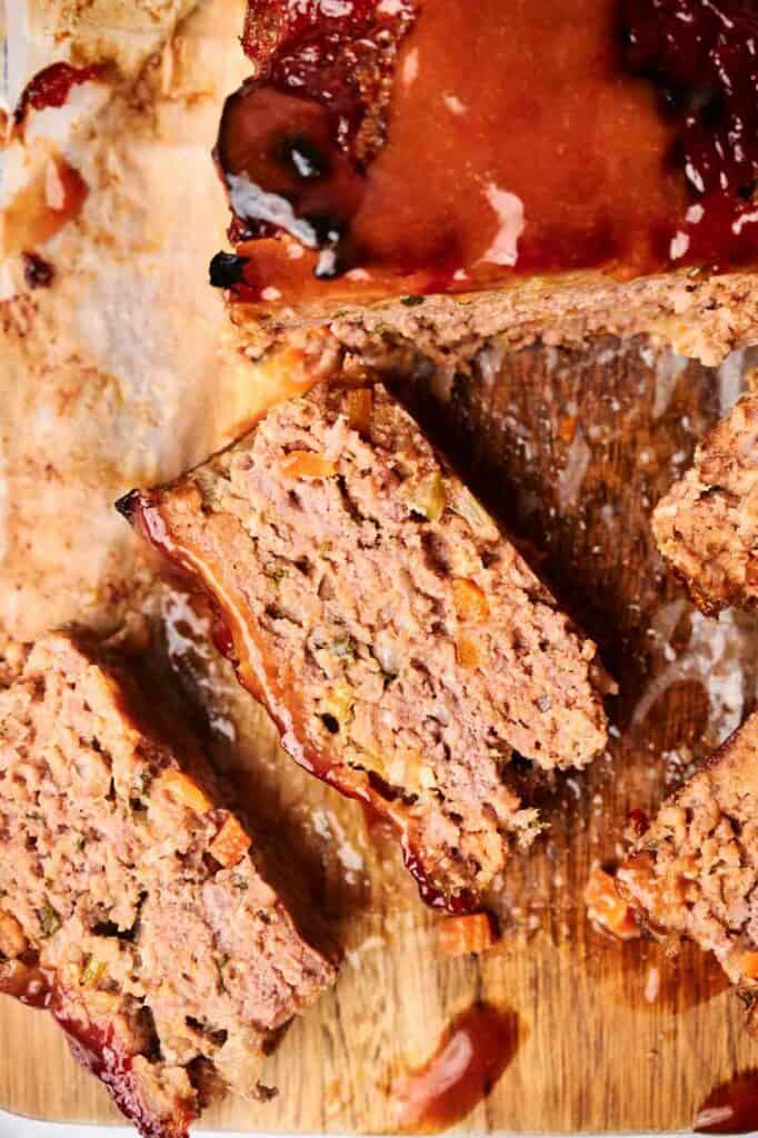 A sliced meatloaf with a glazed topping rests on a wooden cutting board. The meatloaf reveals visible vegetables mixed within, offering a hearty and colorful presentation.