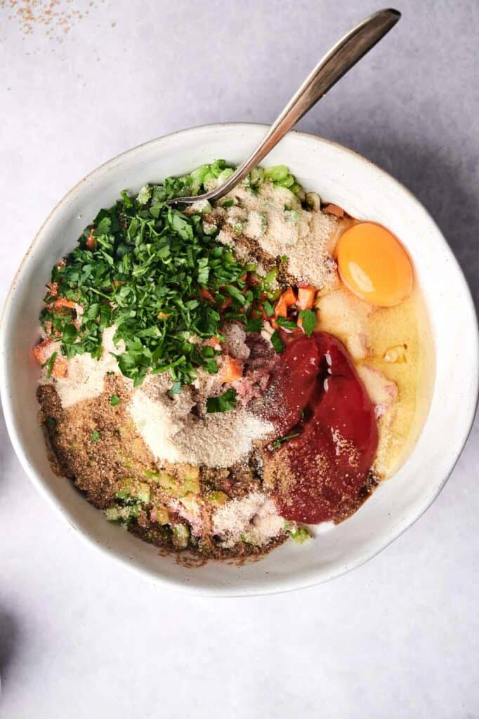 A bowl contains a variety of ingredients including raw egg yolk, chopped herbs, raw meat for meatloaf, breadcrumbs, and vegetables, with a spoon inside.