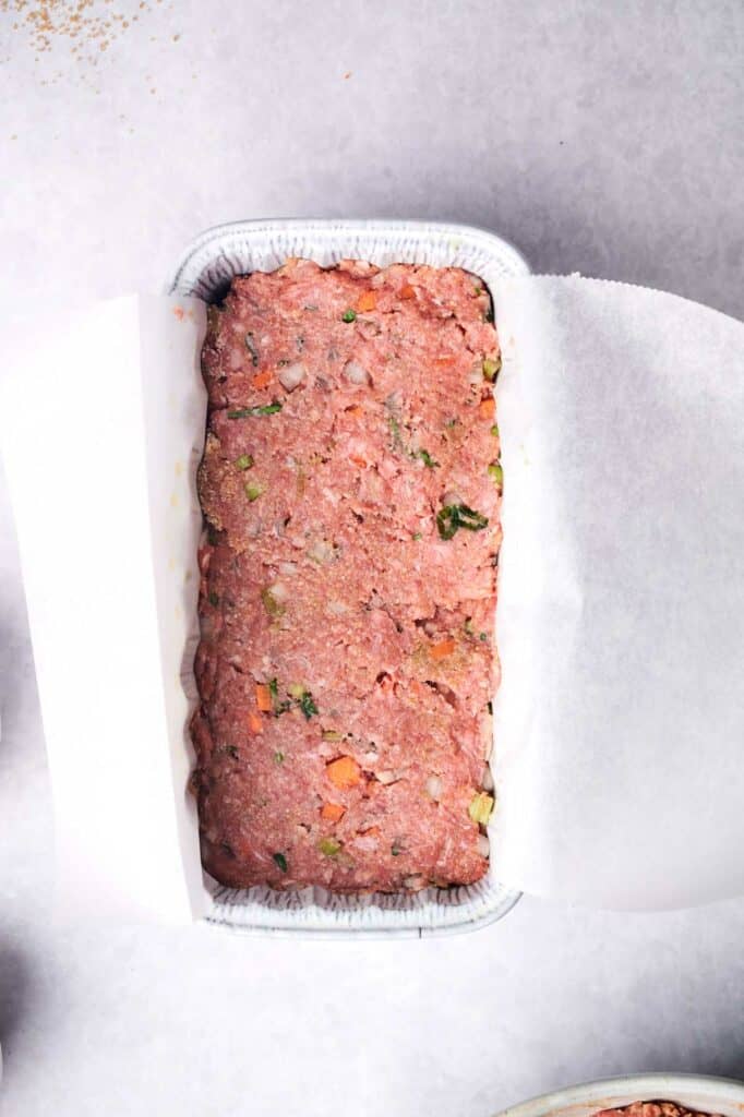 A rectangular metal loaf pan, filled with a meatloaf mixture dotted with visible chunks of vegetables and lined with parchment paper, rests against a light grey surface.