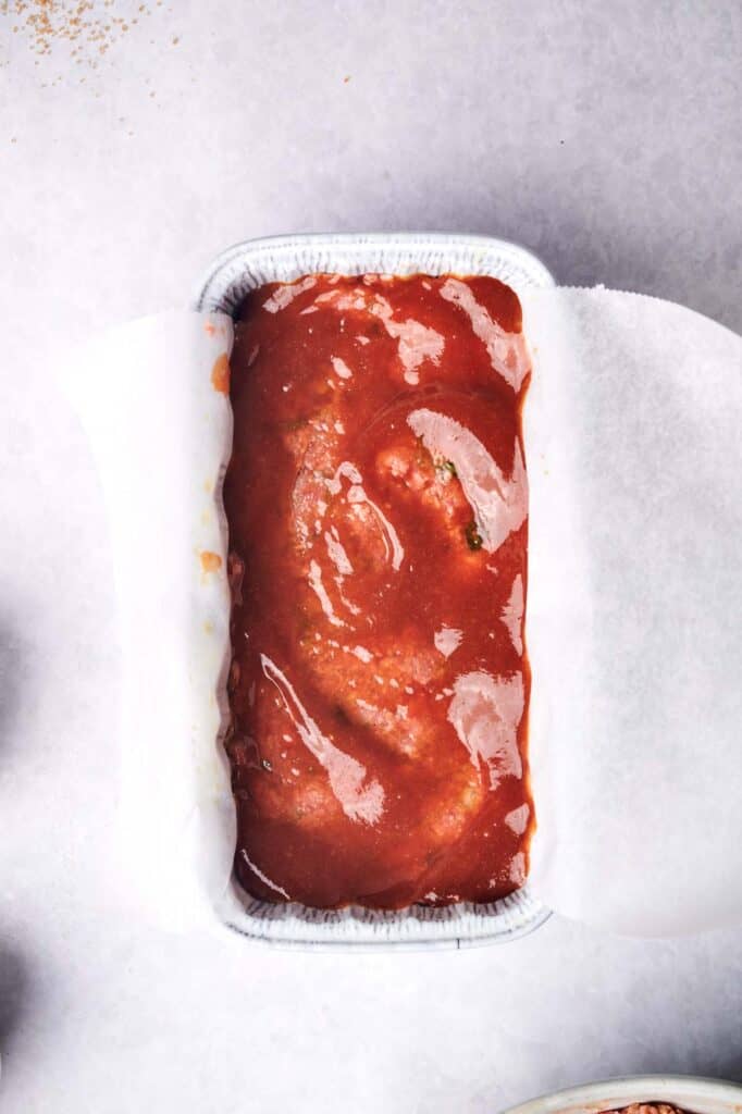 A loaf pan lined with parchment paper contains uncooked meatloaf, generously coated with a rich tomato sauce, shown from above on a light gray surface.