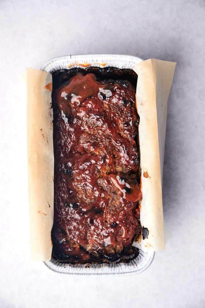 A perfectly seasoned meatloaf, cooked to perfection, sits proudly in a parchment-lined rectangular metal baking dish, its shiny glaze glistening under the light.