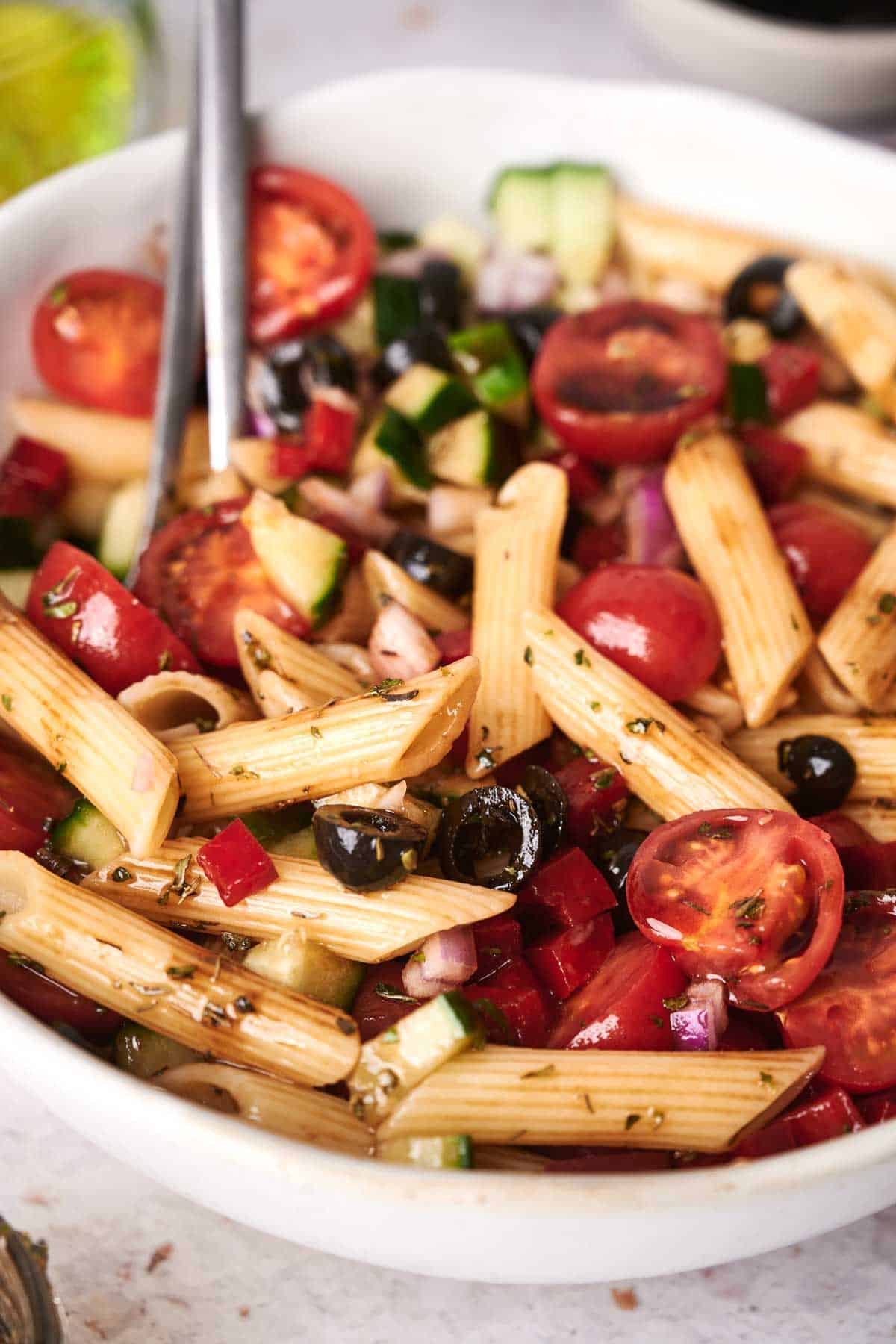 A close-up of a delightful pasta salad in a white bowl, featuring penne, cherry tomatoes, black olives, cucumber, red onion, and red bell pepper. The vibrant dish is beautifully garnished with fresh herbs.