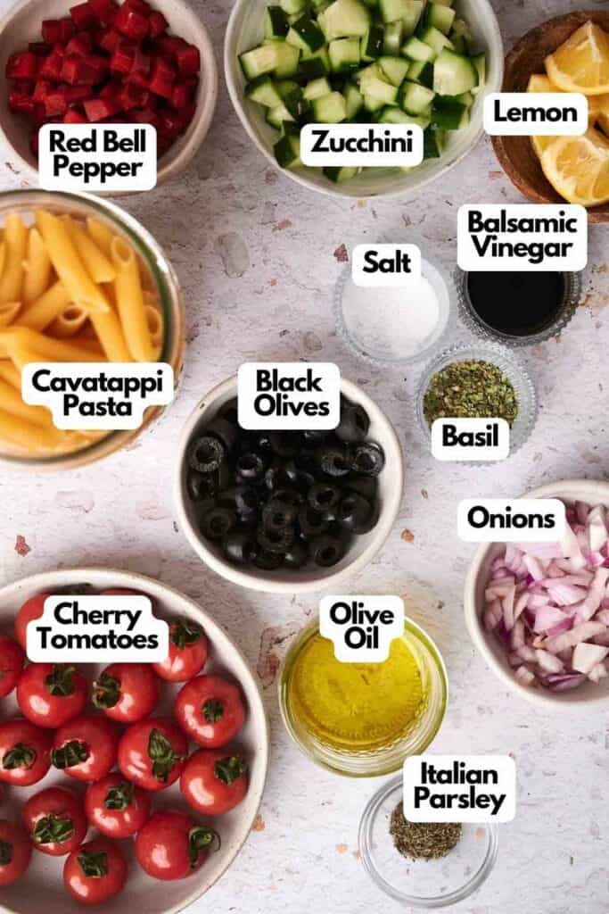 Ingredients for pasta salad laid out in separate bowls: red bell pepper, zucchini, lemon, cavatappi pasta, balsamic vinegar, salt, basil, black olives, onions, cherry tomatoes, olive oil, Italian parsley.