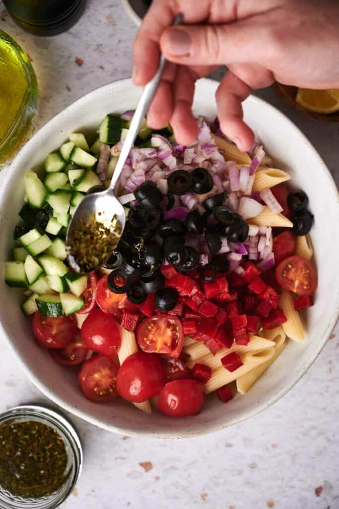 A hand holding a spoon is adding seasoning to a vibrant bowl of pasta salad with tomatoes, cucumbers, red onions, black olives, and red bell peppers.