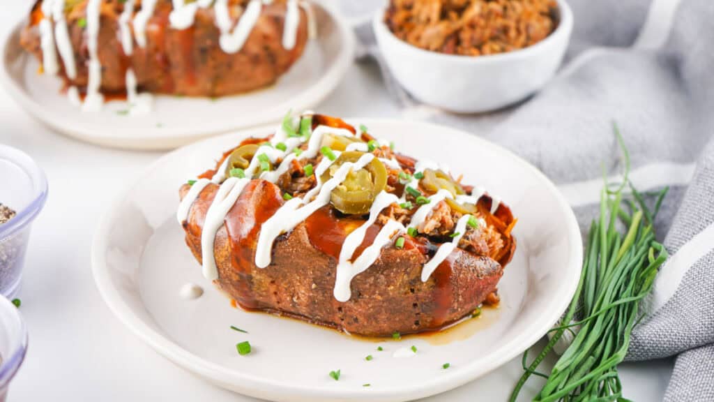 A baked pulled pork potato topped with sliced jalapeños, chives, and a drizzle of white sauce on a white plate. A bowl of rice and additional plate are in the background.
