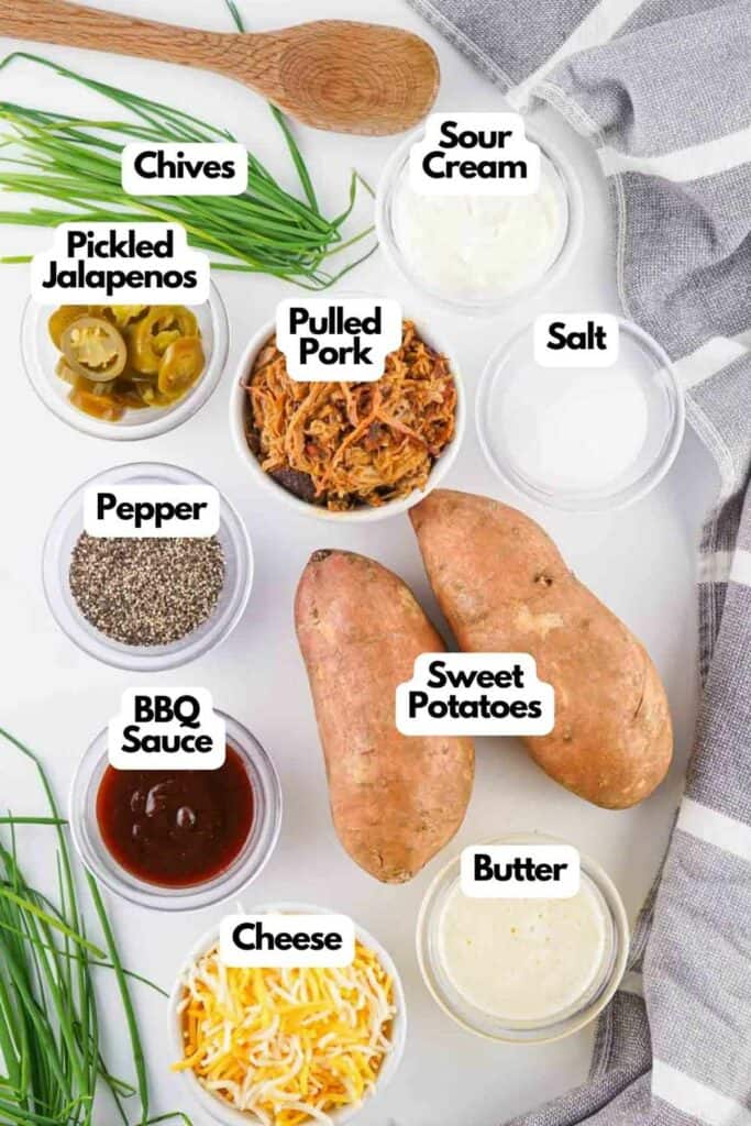 Top view of ingredients for loaded sweet potatoes labeled: chives, sour cream, pickled jalapenos, pulled pork, salt, pepper, sweet potatoes, BBQ sauce, cheese, and butter, on a white surface.