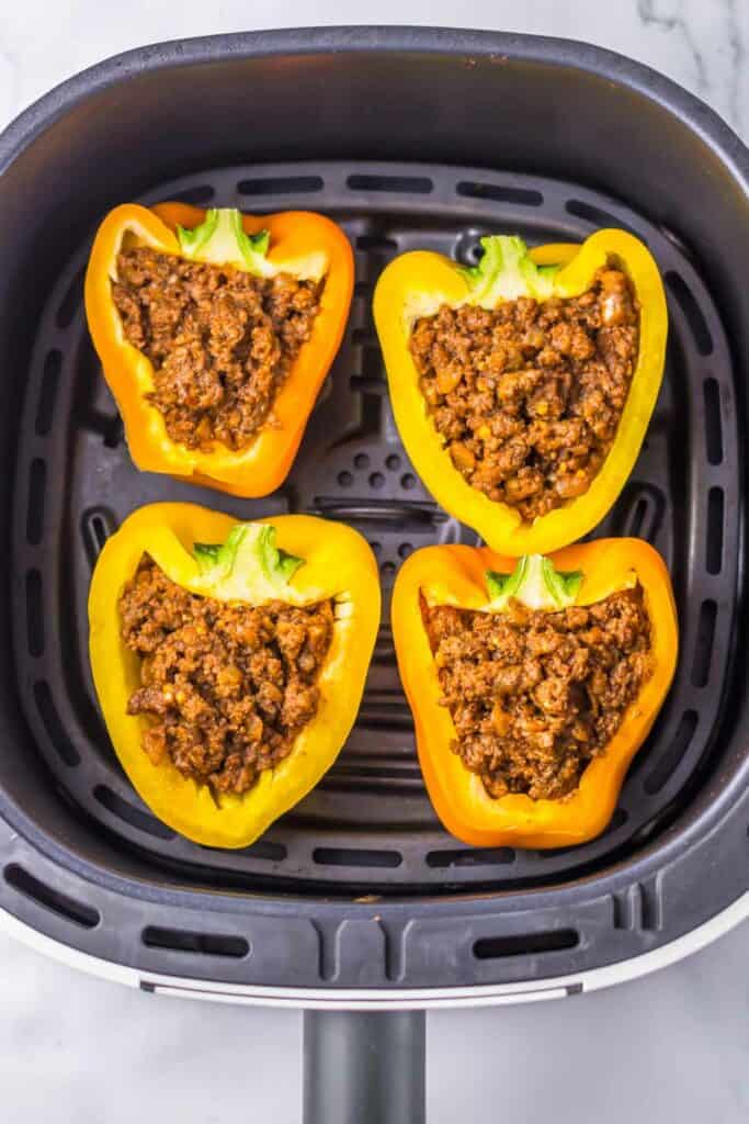 Four halved yellow and orange bell peppers filled with a sloppy joe ground meat mixture are placed inside an air fryer basket.