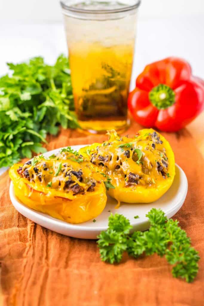 A plate with two sloppy joe stuffed yellow bell peppers topped with melted cheese, garnished with parsley. In the background, there's a whole red bell pepper, fresh parsley, and a glass of iced beverage.