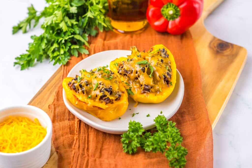 A plate of two sloppy joe stuffed yellow bell peppers topped with melted cheese, garnished with parsley. A cutting board with fresh herbs, a red bell pepper, and a small bowl of shredded cheese are in the background.