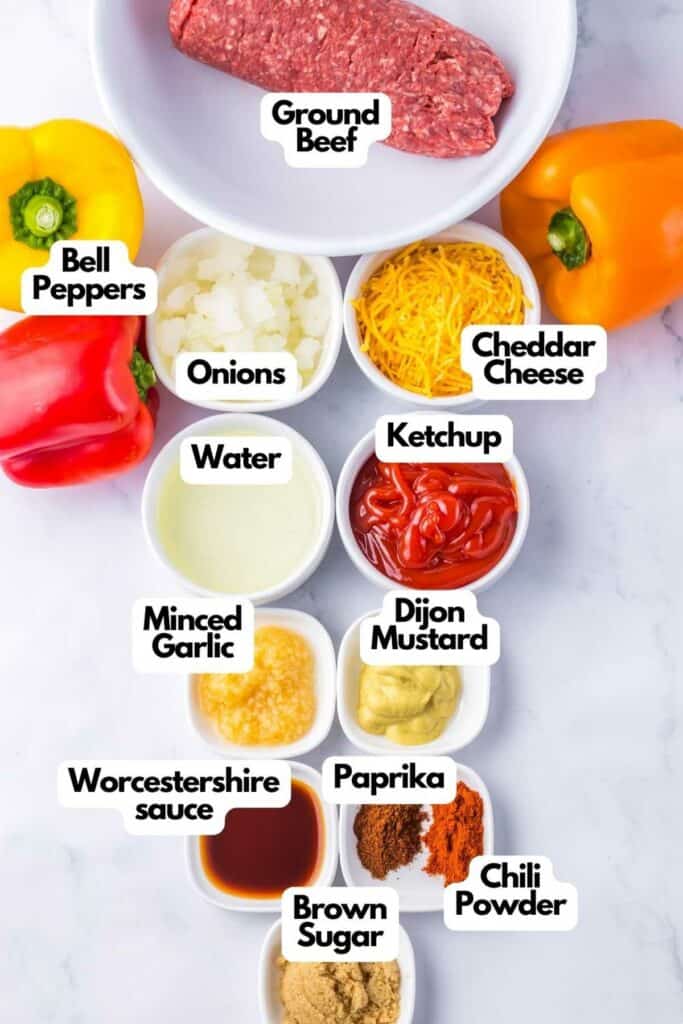 Ingredients for sloppy joe stuffed peppers are displayed on a surface, including ground beef, sliced bell peppers, diced onions, shredded cheddar cheese, water, ketchup, minced garlic, Dijon mustard, Worcestershire sauce, paprika, brown sugar and chili powder.