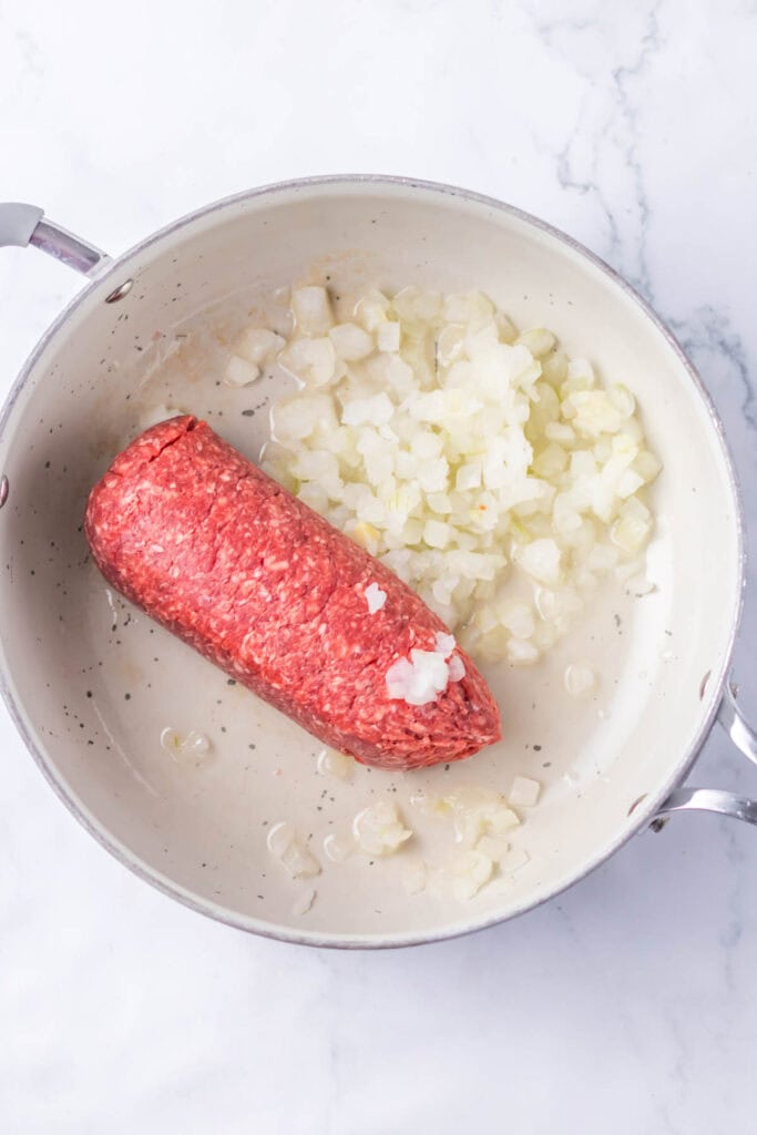 A skillet contains a raw log of ground meat and chopped onions, partially cooked, on a white stove, ready to be transformed into sloppy joe stuffed peppers.