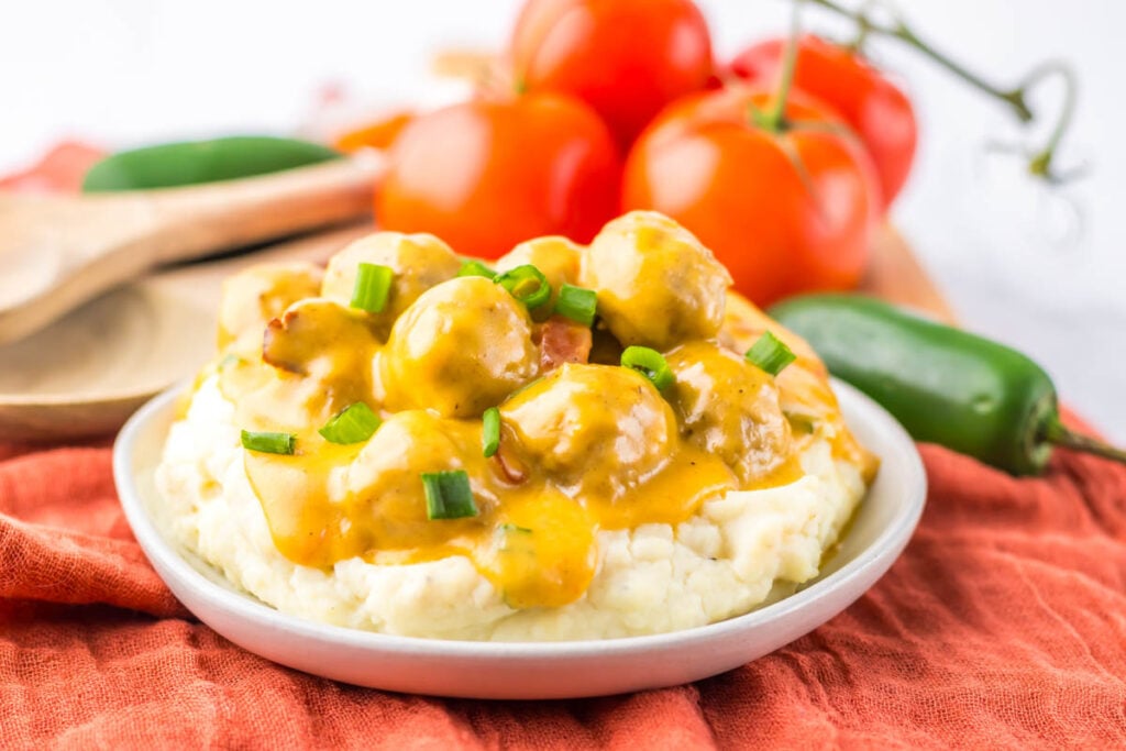Plate of mashed potatoes topped with Slow Cooker Nacho Meatballs in yellow gravy, garnished with chopped green onions; tomatoes and a jalapeño in the background.