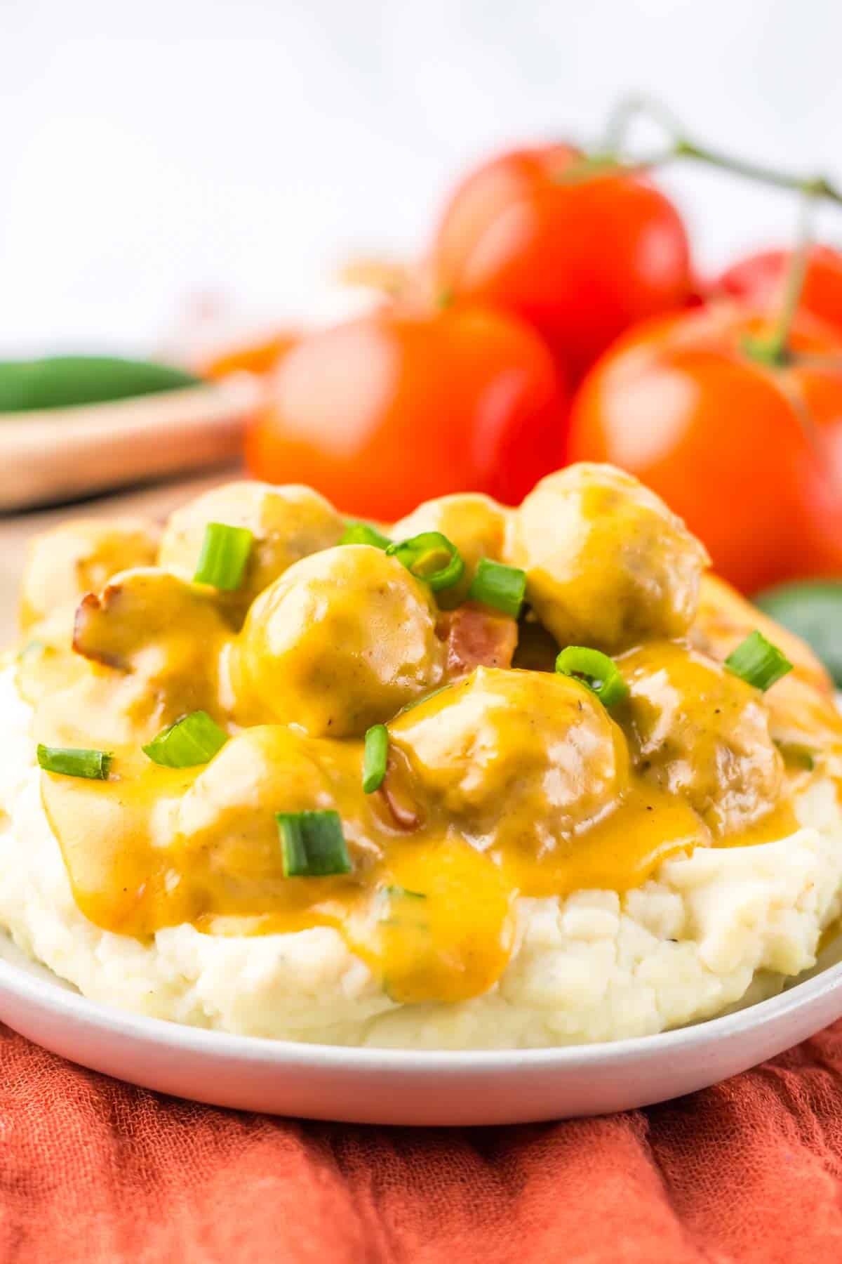 A plate of creamy mashed potatoes topped with mushroom gravy and Slow Cooker Nacho Meatballs, with fresh tomatoes in the background.