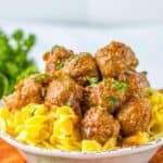 A bowl of slow cooker Salisbury steak meatballs served over egg noodles, garnished with parsley, on a white and orange napkin with fresh parsley on the side.