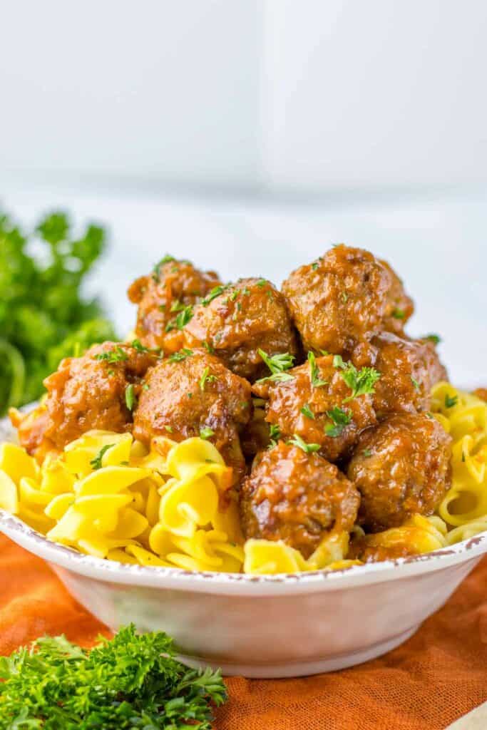 A bowl of slow cooker Sailsbury steak meatballs served over egg noodles, garnished with parsley, on a white and orange napkin with fresh parsley on the side.