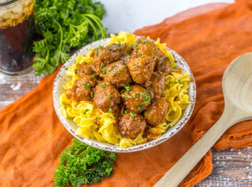 A bowl of egg noodles with slow cooker Salisbury steak meatballs and marinara sauce, garnished with chopped parsley, next to a wooden spoon and a glass of cola on an orange cloth.