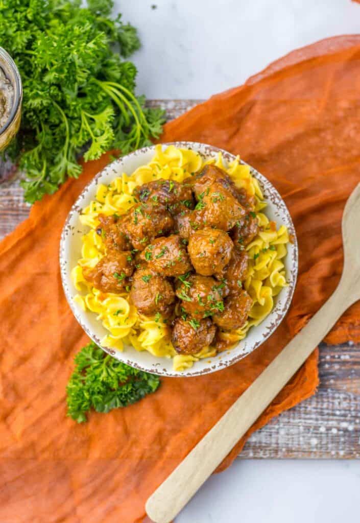A bowl of slow cooker salisbury steak meatballs served over egg noodles, garnished with parsley, placed on a wooden table with a wooden spoon and green herbs nearby.