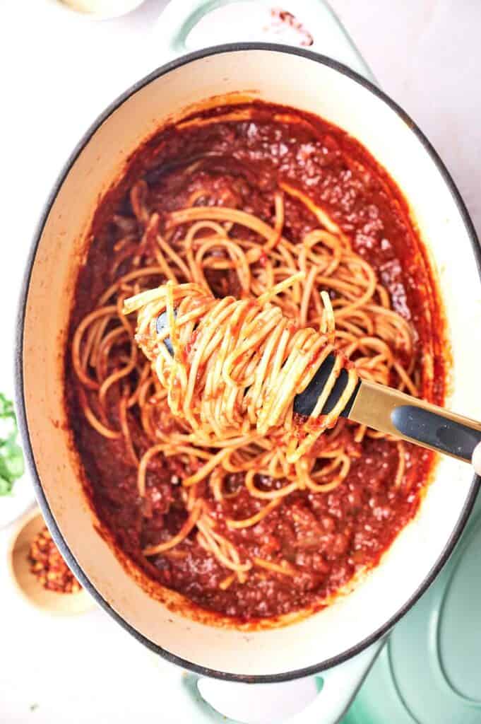 A pot of spaghetti with tomato sauce, with a fork lifting some noodles, on a light-colored surface.