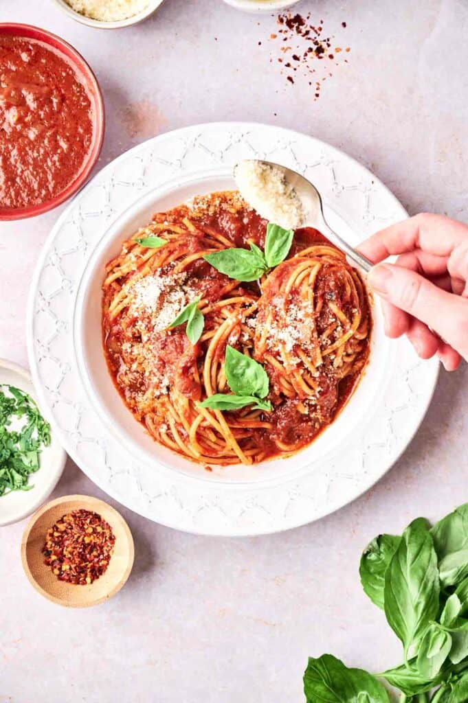 A person's hand sprinkling parmesan cheese on a plate of spaghetti with marinara sauce and fresh basil, surrounded by spices and sauce.