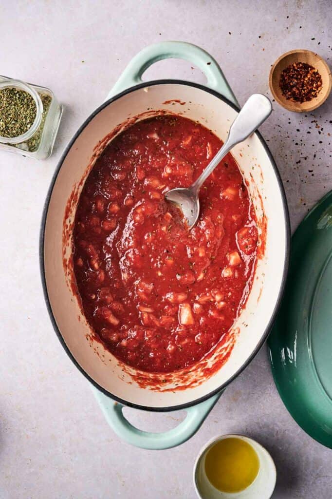 A pot of tomato sauce with a spoon, surrounded by spices and olive oil on a light surface.