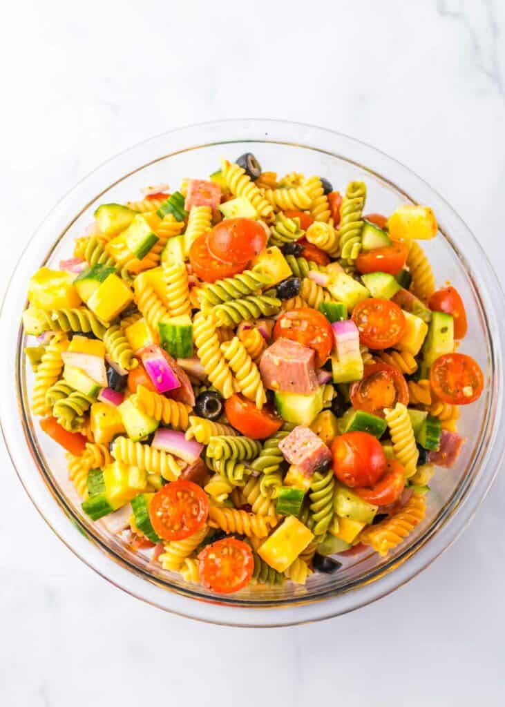 A colorful pasta salad with tomatoes, cucumbers, olives, and diced salami in a glass bowl on a white background.