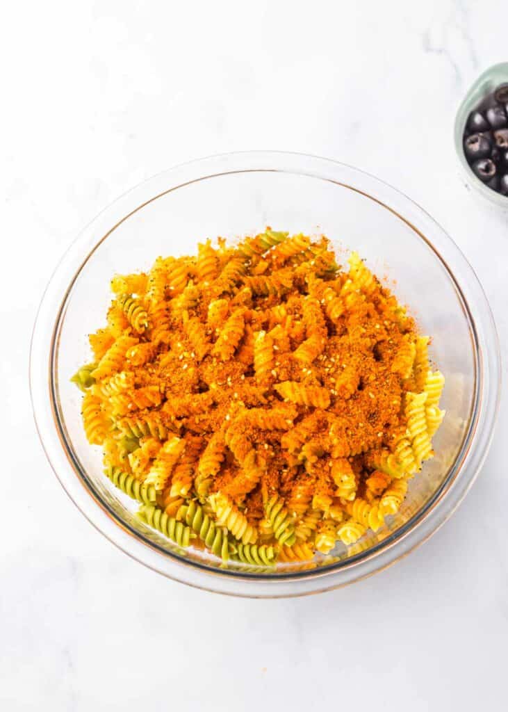 A glass bowl of cooked fusilli pasta topped with orange sauce, on a marble surface with a small bowl of black olives and slices of salami to the side.
