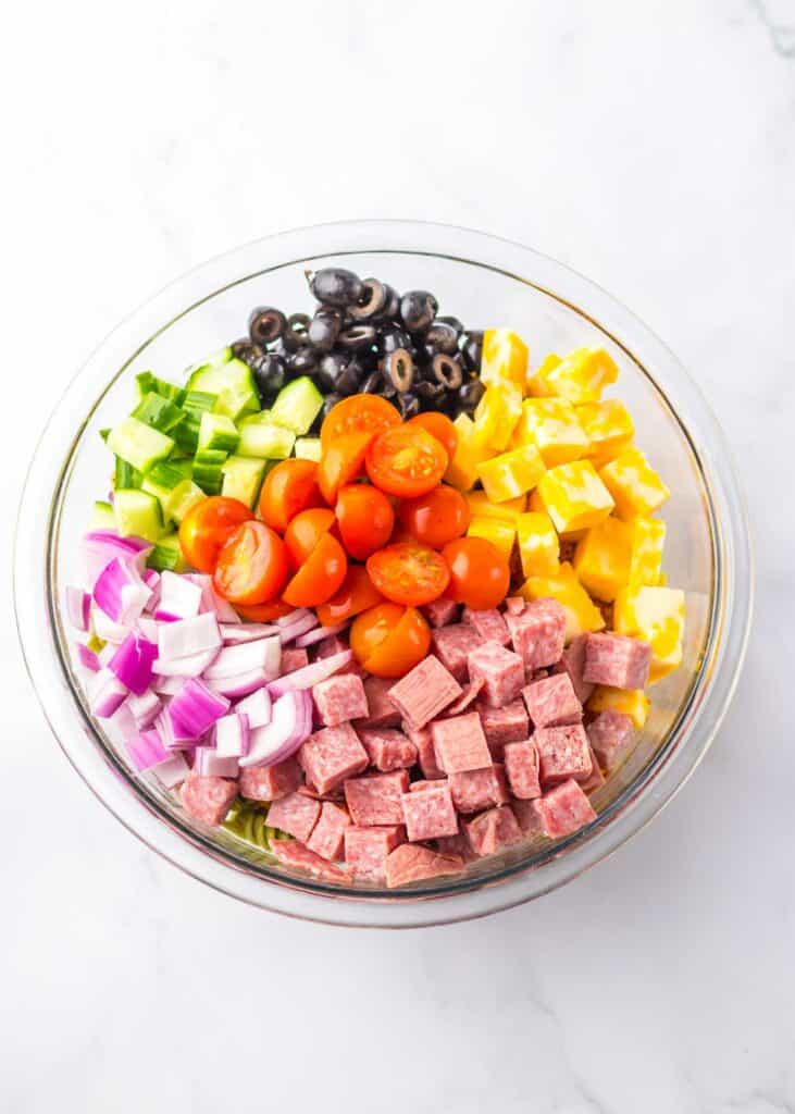A colorful pasta salad in a glass bowl with diced salami, cherry tomatoes, black olives, yellow bell peppers, cucumber, and red onions.
