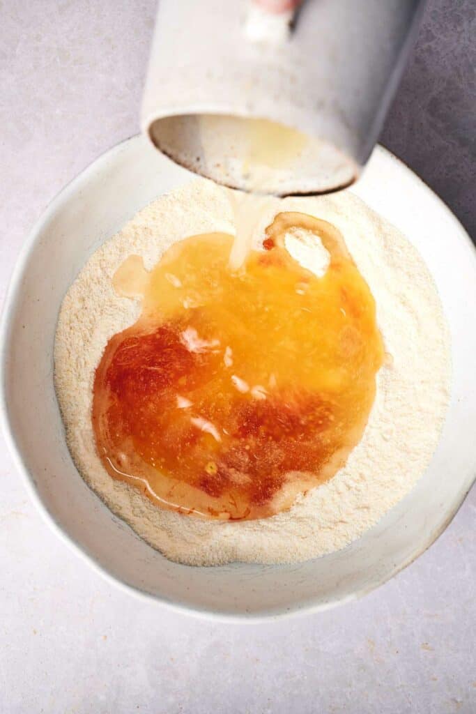 A liquid mixture is being poured from a jug into a bowl containing dry ingredients, likely for baking or preparing tamales.