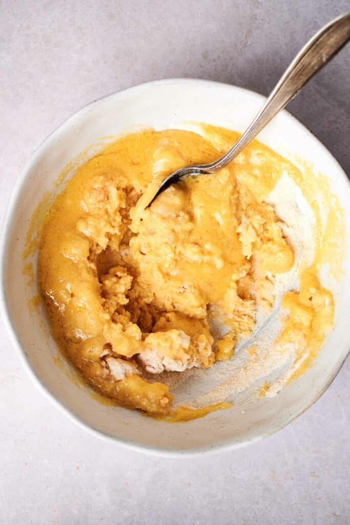 A white bowl contains a thick, yellowish mixture being stirred with a metal spoon, reminiscent of the masa used in making tamales.