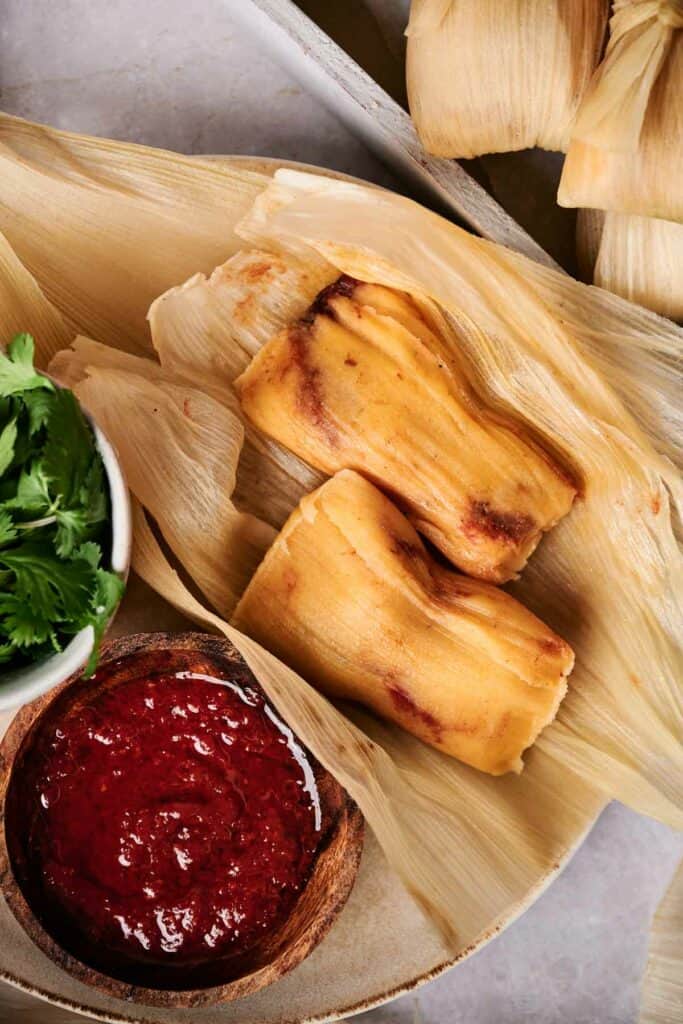 Two tamales on corn husks are next to a bowl of red salsa and a bunch of fresh cilantro, showcasing traditional flavors in every bite.