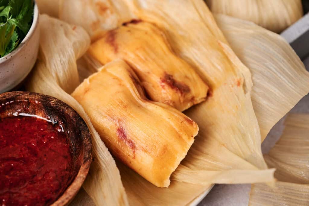 Two tamales lie on a corn husk next to a wooden bowl of tamales' classic red sauce.