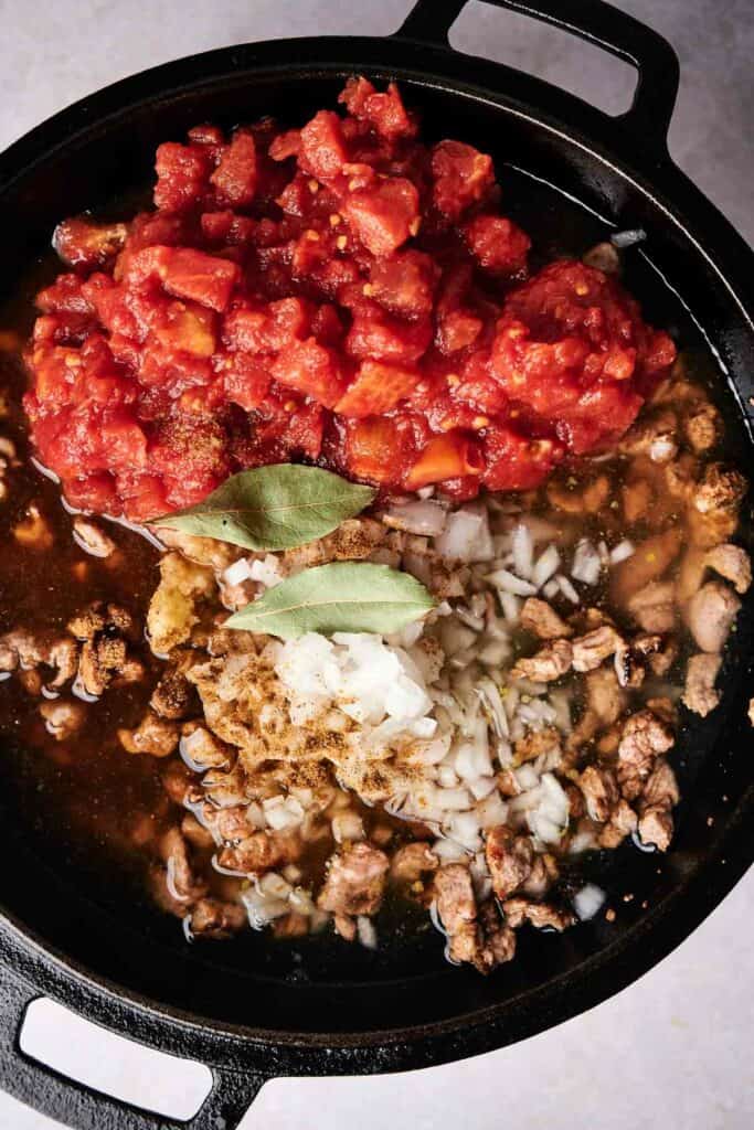 A cast iron skillet with chopped tomatoes, ground meat, diced onions, and spices including bay leaves, all being cooked together for a tamales-inspired filling.