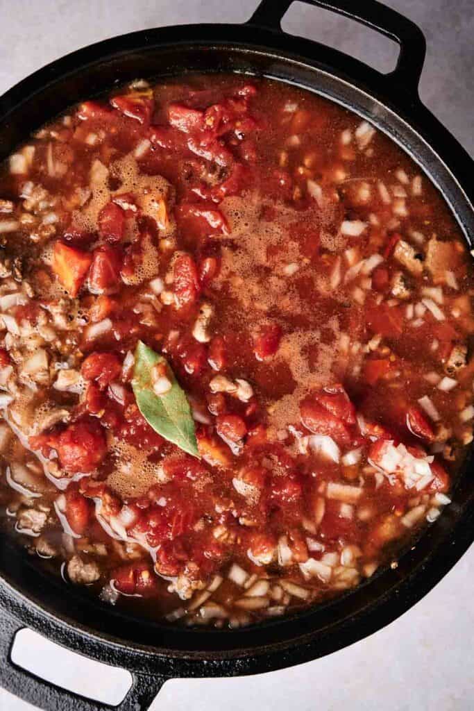 A black pot filled with a chunky stew containing tomatoes, onions, ground meat, and a bay leaf. The stew appears to be simmering, reminiscent of the savory fillings found in traditional tamales.